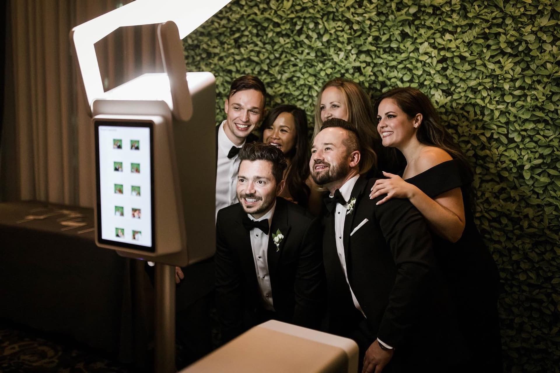 Wedding guests crowded around the photo booth, mid-laugh, in front of a boxwood backdrop
